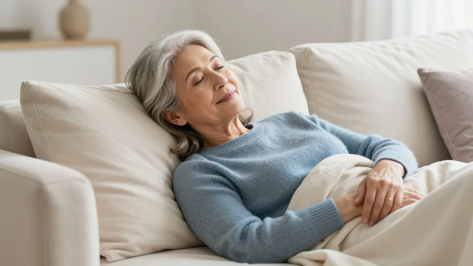 Frau in den Sechzigern mit silbergrauem Haar ruht unter einer leichten Decke auf einem Sofa in einem hellen Wohnzimmer am Nachmittag